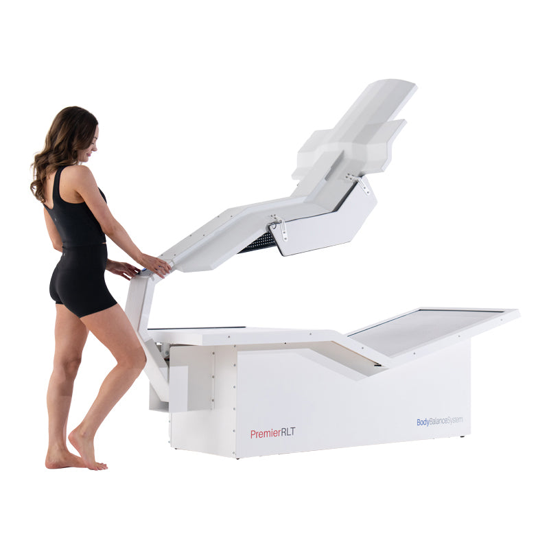Woman demonstrating Body Balance System PremierRLT zero gravity red light therapy bed with canopy open on white background