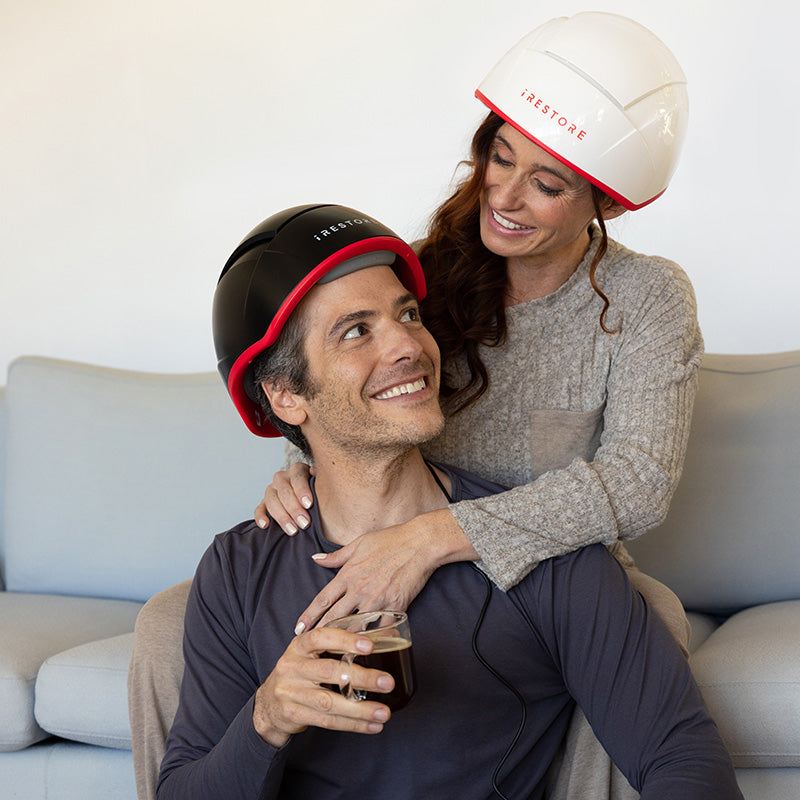 Male and female couple using iRestore Professional Laser Hair Growth System helmets together at home, smiling and enjoying red light therapy session

