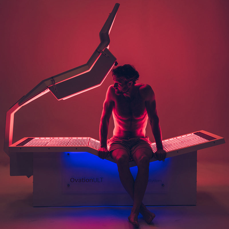 Body Balance System OvationULT Red Light Therapy Bed – front view in dimly lit room with male model seated inside, red light panels illuminated for full-body infrared wellness treatment.