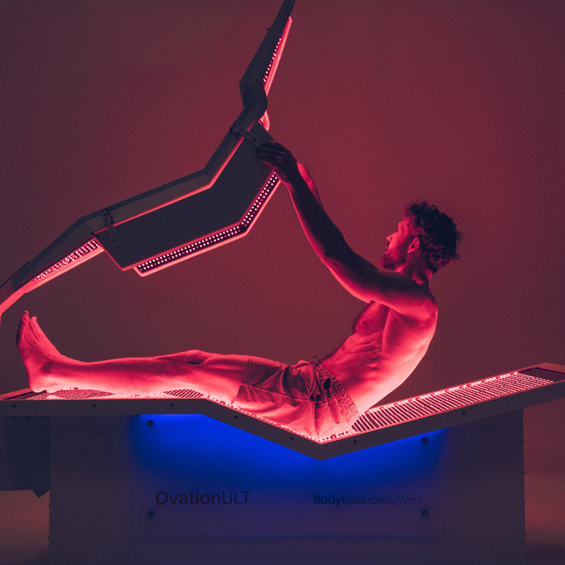 Body Balance System OvationULT Red Light Therapy Bed – male model reclining inside, lowering top canopy in dimly lit room with red light panels illuminated for full-body infrared therapy.