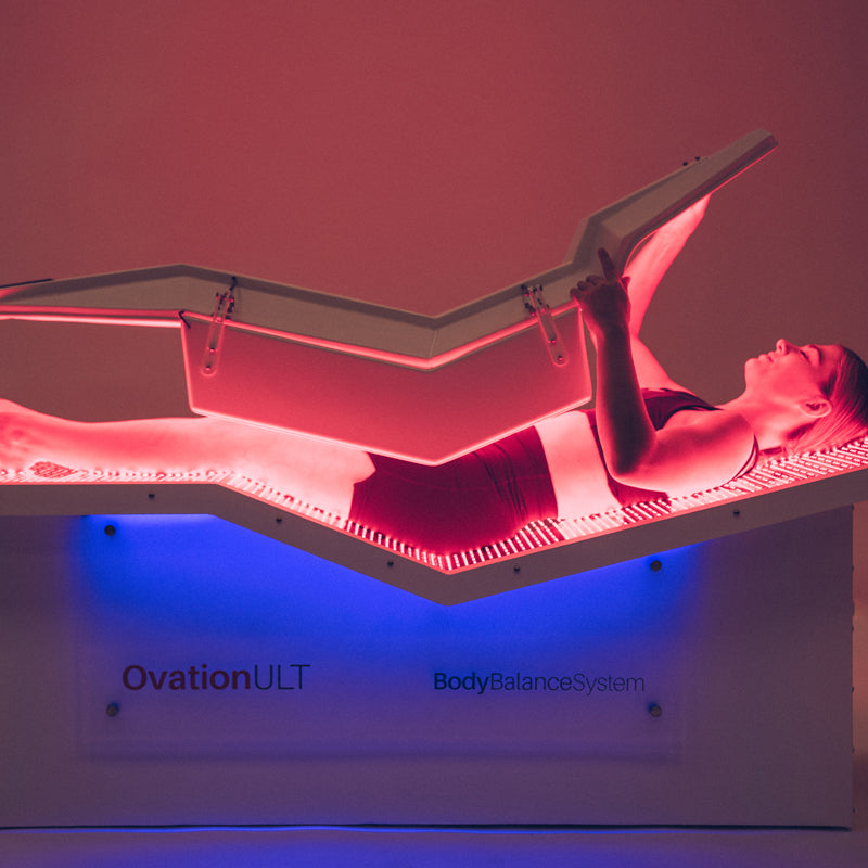 Body Balance System OvationULT Red Light Therapy Bed – female model in dimly lit room closing the top canopy, red light glowing brightly as she begins full-body infrared therapy treatment.
