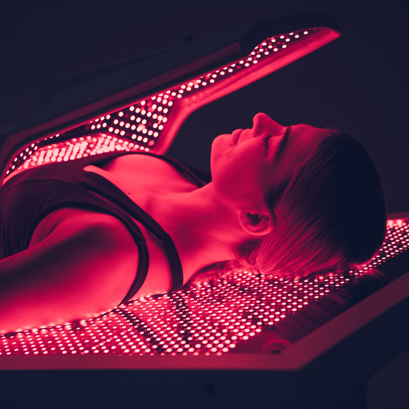 Body Balance System OvationULT Red Light Therapy Bed – close-up of female model’s upper body and face during infrared light treatment in dark room, showcasing red glow and skin rejuvenation benefits.