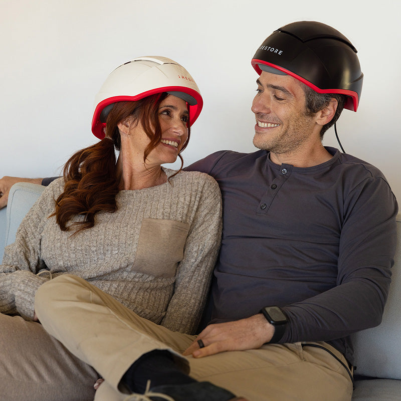 male and female model relaxing on couch both receiving treatment from the iRestore Professional laser hair growth helmet.